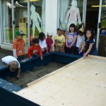 The children stand around as the sand box is uncovered