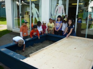 The children stand around as the sand box is uncovered
